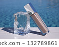 Luxury lip gloss packaging, clear cosmetic product, silver cap, outdoor pool background. Glossy effect, ice cube on white surface, summer beauty concept. Stylish luxury lip gloss. 132789861