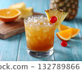 Tropical beverage with pineapple top, orange beside, served with ice in glass on board. Colorful drink offers summer refreshment and exotic appeal. 132789866