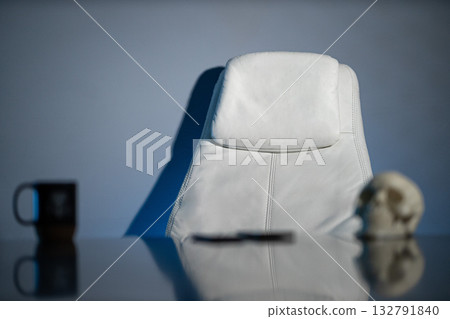 simple white chair sits in a softly lit workspace next to a black mug and a human skull. The scene conveys a minimalist aesthetic with an air of intrigue and contemplation. 132791840