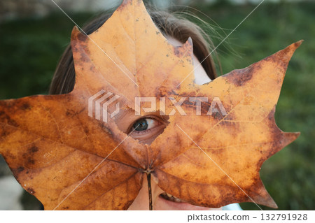 Person playfully hides face behind large autumn leaf with heart-shaped hole showing one eye 132791928