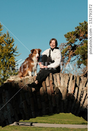 Smiling woman and Australian Shepherd sitting together on an old stone bridge in sunny autumn park 132791972