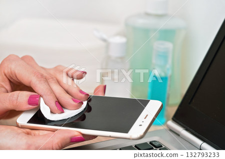 Woman cleaning mobile phone with cotton paper pad, wiping black screen, closeup detail. Blurred bottles and laptop keyboard in background 132793233
