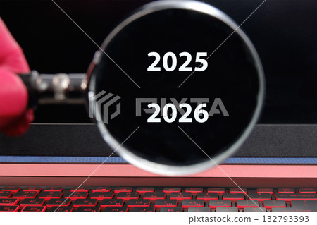 A hand in a pink glove holds a magnifying glass over the screen of a laptop, revealing the numbers 2025 and 2026. A red keyboard sits below the screen 132793393