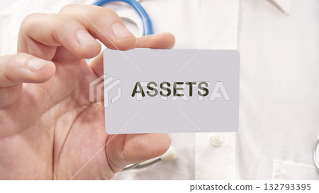 Doctor wearing a white coat holds a card that reads ASSETS, implying financial responsibility in medical care and value in personal wellbeing in a hospital environment 132793395