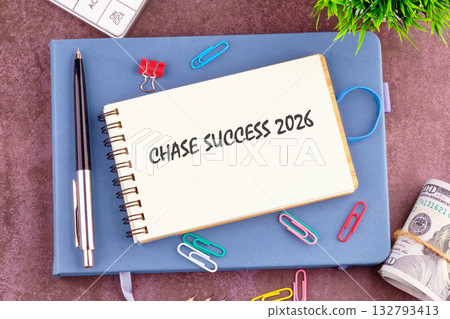 A notebook with the words CHASE SUCCESS 2026 rests on a blue journal. A pen, paper clips, a calculator, some cash, and a small plant are visible A notebook with the words CHASE SUCCESS 2026 rests on a blue journal. A pen, paper clips, a calculator, some cash, and a small plant are visible 132793413
