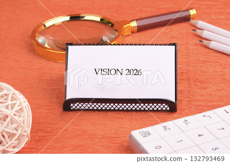 on an orange desk, a white card reading Vision 2026 sits near a magnifying glass, pencils, a calculator, and a decorative woven ball, suggesting future planning on an orange desk, a white card reading Vision 2026 sits near a magnifying glass, pencils, a calculator, and a decorative woven ball, suggesting future planning 132793469