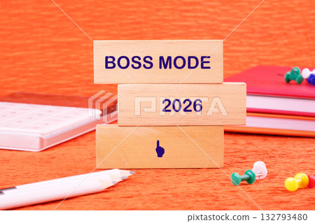 Three wooden blocks stacked show BOSS MODE on top and 2026 in the middle. A pencil, notebook and push pins rest on a textured orange background 132793480