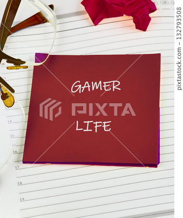 Desk planner lies open, featuring a crimson card with GAMER LIFE in bold white letters, accompanied by glasses, a crumpled paper flower, and numbered lines on the page Desk planner lies open, featuring a crimson card with GAMER LIFE in bold white letters, accompanied by glasses, a crumpled paper flower, and numbered lines on the page 132793508