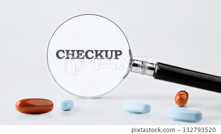A magnifying glass is positioned over the word CHECKUP, with several capsules and pills of various colors placed around it on a white surface A magnifying glass is positioned over the word CHECKUP, with several capsules and pills of various colors placed around it on a white surface 132793520
