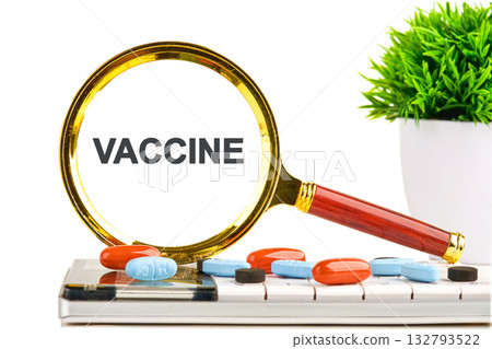 A magnifying glass highlights the word VACCINE on what appears to be a computer screen. Pills are scattered on a keyboard next to a potted plant 132793522