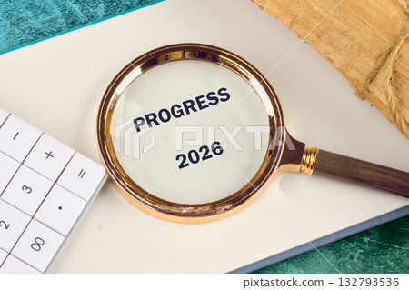 Magnifying glass focuses on the word PROGRESS and the year 2026 sitting on a notebook. Beside it, there is a calculator and piece of wood rolled up with twine 132793536