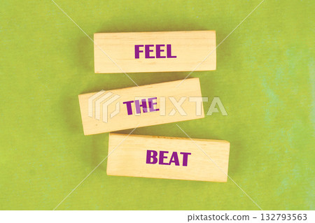 Three light wooden blocks with the words FEEL THE BEAT in purple font are staggered on a bright green background, urging viewers to connect with the rhythm and energy of sound 132793563