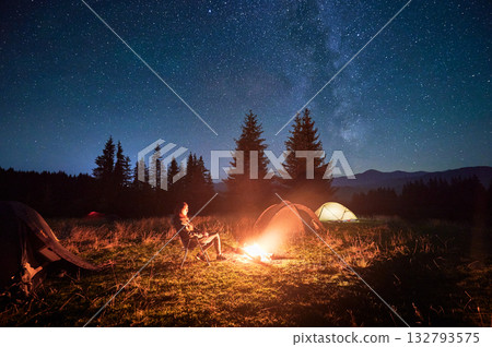 Hiker woman relaxes by campfire, illuminated by its warm glow, with tent nearby under stunning, star-filled sky. Milky Way stretches overhead, adding magical touch to serene mountain setting. 132793575