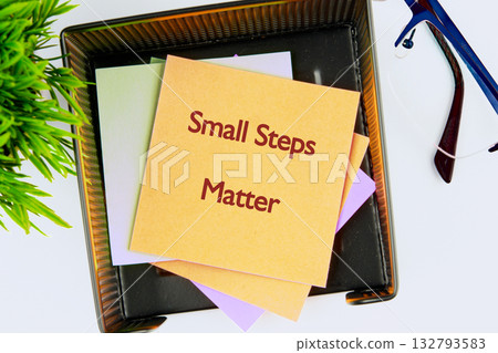 A stack of colorful sticky notes sitting in a black tray on a white desk. A top note reads Small Steps Matter. Glasses and a potted plant sit nearby A stack of colorful sticky notes sitting in a black tray on a white desk. A top note reads Small Steps Matter. Glasses and a potted plant sit nearby 132793583