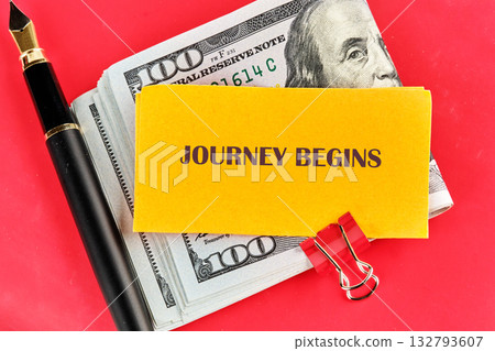 A stack of United States currency, a yellow notepad with the words Journey Begins, a red binder clip, and a black fountain pen lay on a red surface 132793607
