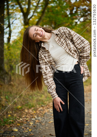 A young woman with long hair smiles brightly while wearing a plaid shirt and black pants. She stands on a path surrounded by colorful autumn trees in the park. A young woman with long hair smiles brightly while wearing a plaid shirt and black pants. She stands on a path surrounded by colorful autumn trees in the park. 132793630
