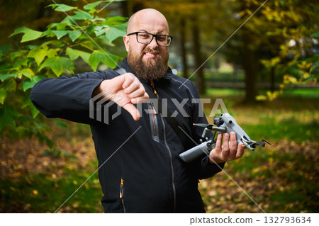 In a vibrant park filled with golden leaves, a man expresses frustration while holding a drone. His thumbs down signal highlights a setback in his outdoor adventure. In a vibrant park filled with golden leaves, a man expresses frustration while holding a drone. His thumbs down signal highlights a setback in his outdoor adventure. 132793634