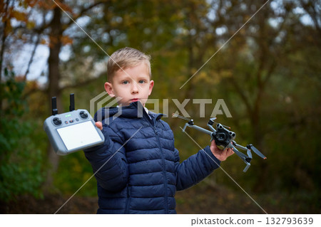 A boy stands in a vibrant autumn setting, confidently holding a drone in one hand and a controller in the other. Leaves in various colors surround him, highlighting the season. 132793639