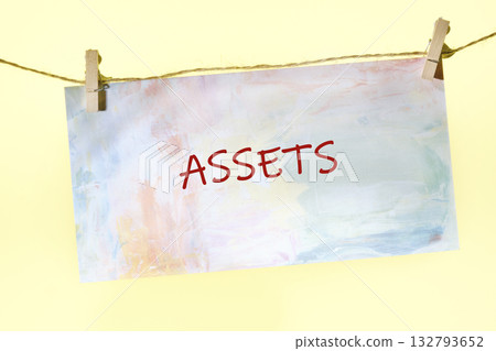 A colorful painting with the word ASSETS written in red hangs from a string with wooden clothespins. The background is a soft, sunny yellow color, creating a hopeful mood A colorful painting with the word ASSETS written in red hangs from a string with wooden clothespins. The background is a soft, sunny yellow color, creating a hopeful mood 132793652
