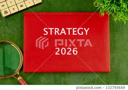 A red book titled Strategy 2026 sits on a textured green surface. Theres a calculator, magnifying glass, and a green plant also in view A red book titled Strategy 2026 sits on a textured green surface. Theres a calculator, magnifying glass, and a green plant also in view 132793689