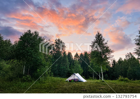 White tourist tent with camping gear in grassy clearing against backdrop of dense trees. Sky glows with vibrant hues of orange and pink as sun sets, casting serene ambiance over peaceful camping site. 132793754