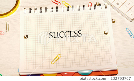 A grid notebook lays open on a desk, prominently displaying the word success in bold letters. Colorful paper clips scatter around, adding a creative touch to the workspace A grid notebook lays open on a desk, prominently displaying the word success in bold letters. Colorful paper clips scatter around, adding a creative touch to the workspace 132793767