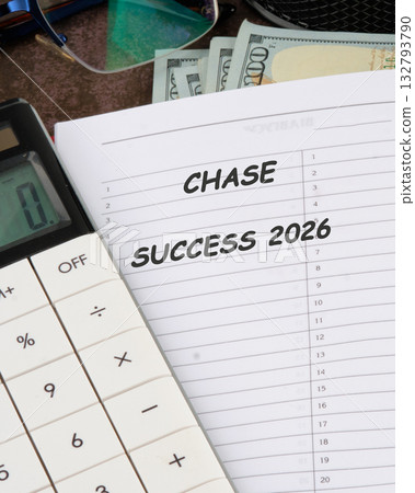 A calculator sits beside a notepad labeled with chase success 2026, surrounded by cash and office supplies, symbolizing the pursuit of financial goals and careful planning 132793790