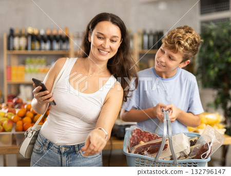 Mom chooses products from a list on her smartphone. Teenage son helps carry shopping cart in supermarket 132796147