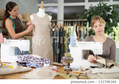 Fashion designer working at sewing machine in studio with teammate 132796168