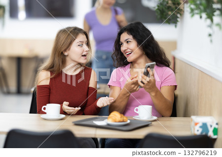 Two positive young girls chatting over coffee and croissant while scrolling smartphone Two positive young girls chatting over coffee and croissant while scrolling smartphone 132796219