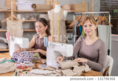 Two positive female dressmakers working with sewing-machine in tailor's workshop Two positive female dressmakers working with sewing-machine in tailor's workshop 132796368