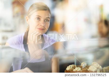 Positive young woman offering pastries standing behind glass display Positive young woman offering pastries standing behind glass display 132796525
