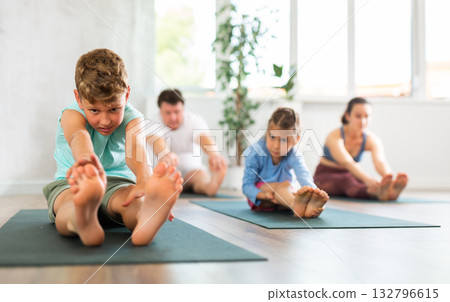 Preschool child performs back stretching pose, Paschimottasana. Family does hatha yoga in background 132796615
