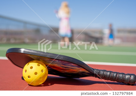 Pickleball paddle and ball resting on a court while players compete in the background under a clear blue sky 132797984