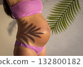 Pregnancy concept. Pregnant woman in pink bikini with belly exposed, tropical leaves in foreground. 132800182