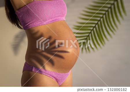 Pregnancy concept. Pregnant woman in pink bikini with belly exposed, tropical leaves in foreground. 132800182