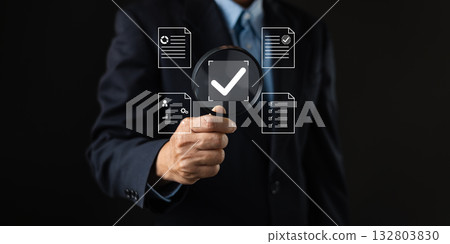 A businessman holds a magnifying glass over a digital checkmark surrounded by document icons, symbolizing document review, quality control, approval process, and data verification in business. 132803830