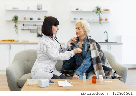 A female doctor uses a stethoscope to listen to the chest of an elderly woman sitting on a couch at home. There are medicines on the table. 132805561