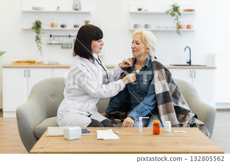 A female doctor uses a stethoscope to listen to the chest of an elderly woman sitting on a sofa during a home visit. The patient is wrapped in a blanket. 132805562