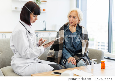 A female doctor examines an elderly patient at home, using a tablet to review medical information. The patient appears to be unwell. 132805563