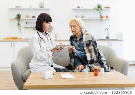 A female doctor in a white coat consults with an elderly patient wrapped in a blanket, discussing health concerns. 132805568