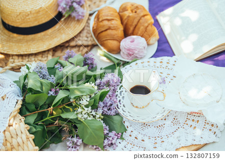 Charming spring picnic setup featuring fresh lilac flowers, a cup of coffee in fine china, croissants, marshmallow dessert, soft cheese, and an open book on a crocheted lace cloth. 132807159