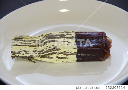 A bite-sized piece of Shimizu Yokan, with its individual wrapper peeled off and half of the contents squeezed out, is placed horizontally on a white plate. A bite-sized piece of Shimizu Yokan, with its individual wrapper peeled off and half of the contents squeezed out, is placed horizontally on a white plate. 132807614