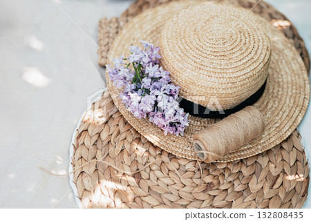 Elegant flat lay with straw hat decorated with lilac flowers and twine placed on natural woven mat. Soft light and neutral tones create calm and summery mood, perfect for lifestyle or nature themes. 132808435