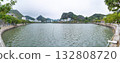 A serene panorama of a calm lake surrounded by lush greenery and a backdrop of mountains in Vietnam. The tranquil water reflects the overcast sky. 132808720