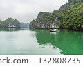 A serene boat floats on the tranquil waters of Ha Long Bay, Vietnam, surrounded by lush limestone karsts. The emerald water mirrors the overcast sky. 132808735