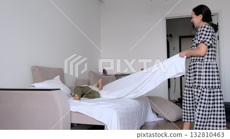 A playful scene shows a woman in a checkered dress making the bed as her boy grins beneath rising linen, framed wide and natural. This daily chore builds teamwork, independence, and calm at home. 132810463