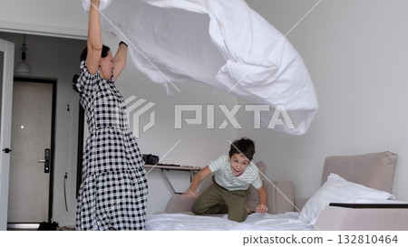 A mother playfully shakes a blanket while her son eagerly jumps on the bed. This joyful scene captures family bonding and the simple pleasures of spending time together at home. A mother playfully shakes a blanket while her son eagerly jumps on the bed. This joyful scene captures family bonding and the simple pleasures of spending time together at home. 132810464