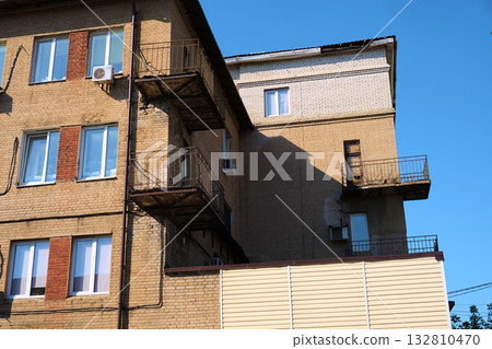 Old residential brick building with deteriorating balconies, cracked plaster, and visible patch repairs on the facade. The photo reflects structural wear and lack of maintenance, suitable for Old residential brick building with deteriorating balconies, cracked plaster, and visible patch repairs on the facade. The photo reflects structural wear and lack of maintenance, suitable for 132810470