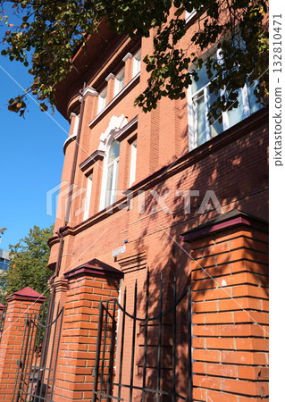 Facade of an old red brick building with arched windows and decorative white accents, partially shaded by trees and sunlight reflections. The image is suitable for topics about building restoration Facade of an old red brick building with arched windows and decorative white accents, partially shaded by trees and sunlight reflections. The image is suitable for topics about building restoration 132810471